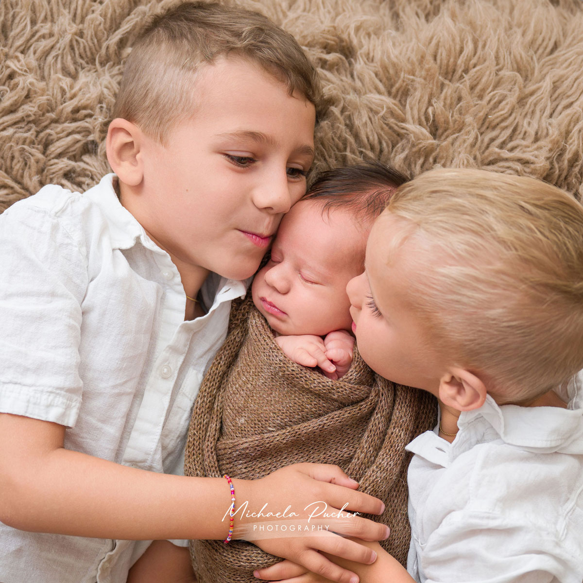 Zwei ältere Geschwister kuscheln sich von beiden Seiten an ihr schlafendes neugeborenes Geschwisterchen, das in braune Tücher gewickelt ist.
